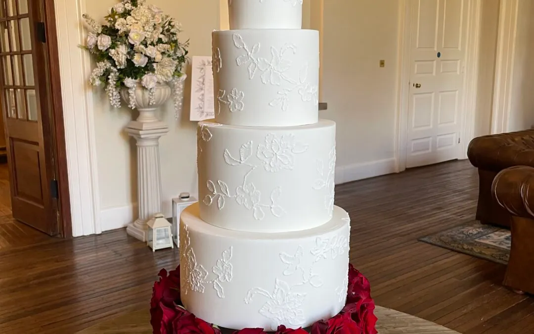 Four-tier white wedding cake with delicate brush embriodery detailing, decorated with deep red roses on the top and around the base, displayed on a glass stand in an elegant venue.